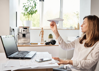 Woman procrastinates with paper airplane at desk.