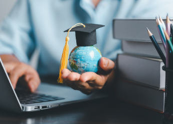 Hand holding globe with grad cap while using laptop